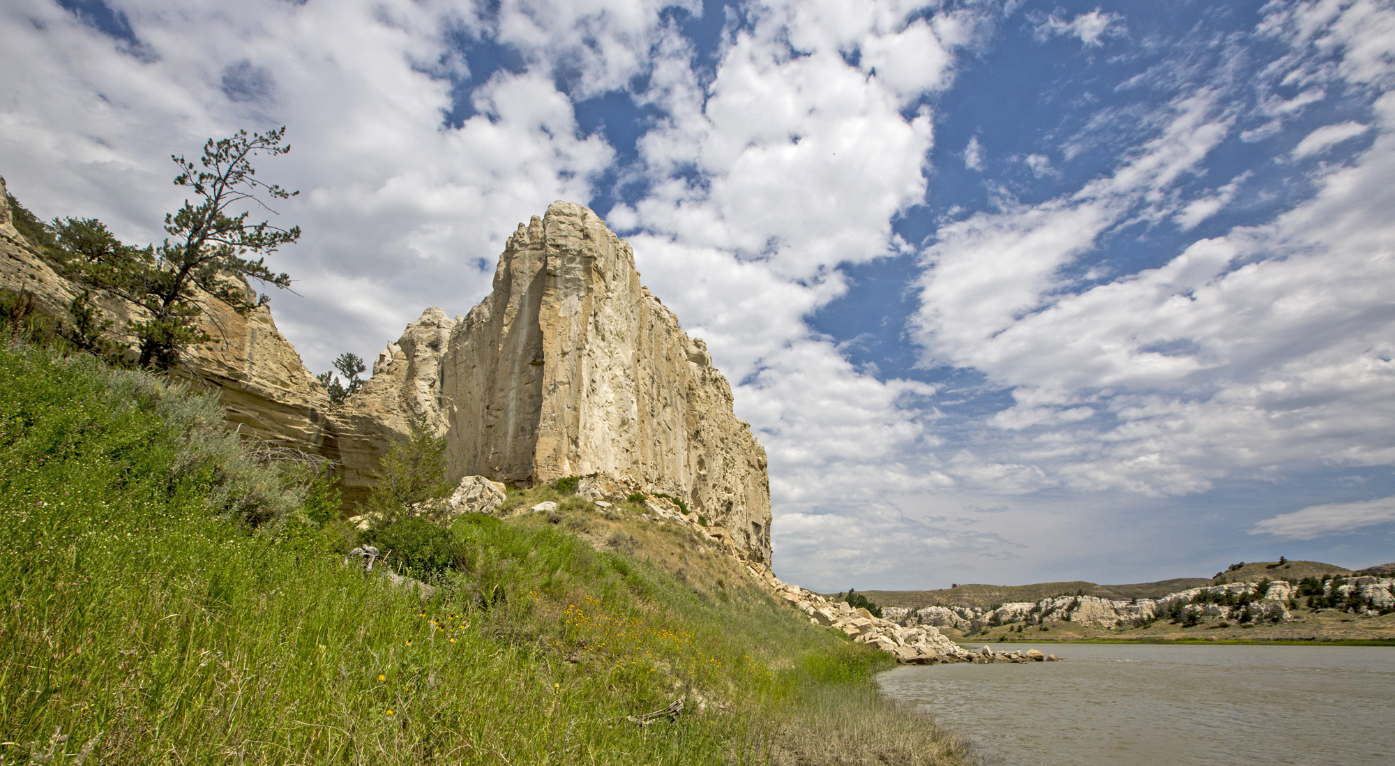 Photo. Daytime; sunny, partly-cloudy, blue skies above towering white, sandstone cliffs. The Missouri river flows below the cliffs, blue-gray water; grassy, sloped shoreline on the left; rocky, cliff-like shoreline on the right.